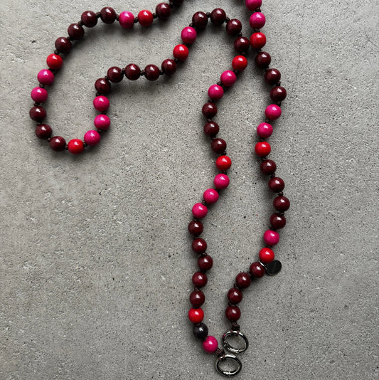 A beaded necklace made of alternating dark burgundy, bright pink, and red round beads, laid out on a textured gray concrete surface. It features two silver metal rings at the bottom.