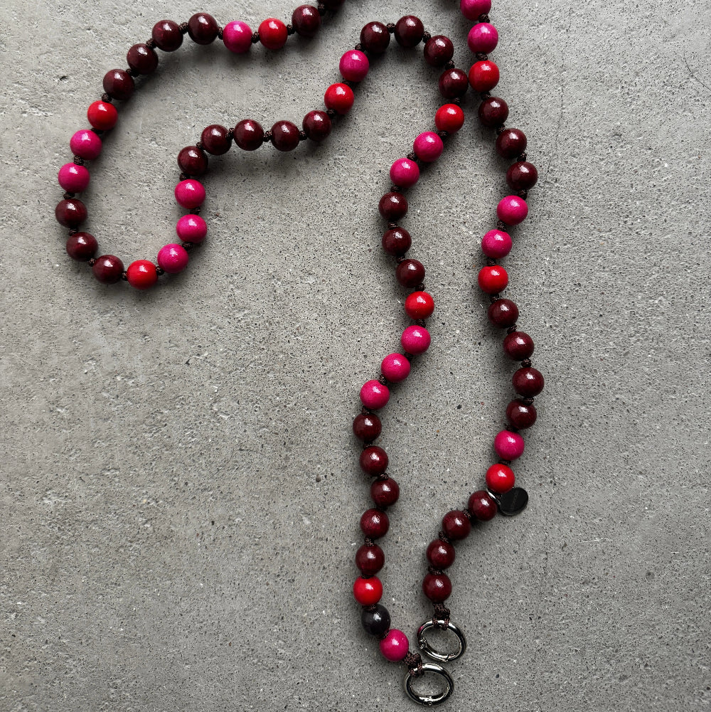 A beaded necklace made of alternating dark burgundy, bright pink, and red round beads, laid out on a textured gray concrete surface. It features two silver metal rings at the bottom.