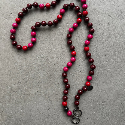 A beaded necklace made of alternating dark burgundy, bright pink, and red round beads, laid out on a textured gray concrete surface. It features two silver metal rings at the bottom.