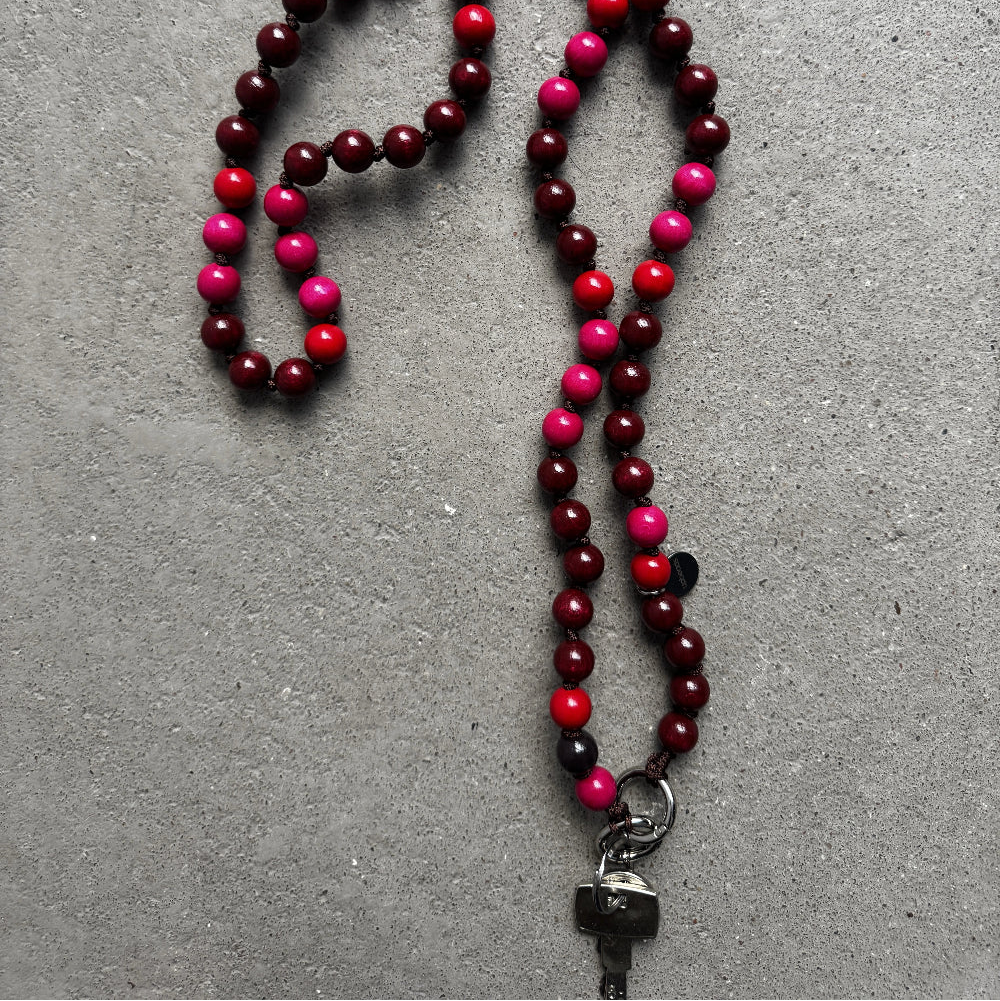 A beaded upbeads crossbody chain with alternating bordeaux red, bright red, and hot pink wooden beads, featuring a silver metal carabiner used as a keychain, laid out on a textured gray surface.