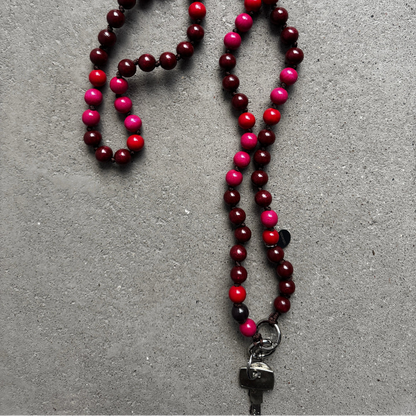 A beaded upbeads crossbody chain with alternating bordeaux red, bright red, and hot pink wooden beads, featuring a silver metal carabiner used as a keychain, laid out on a textured gray surface.