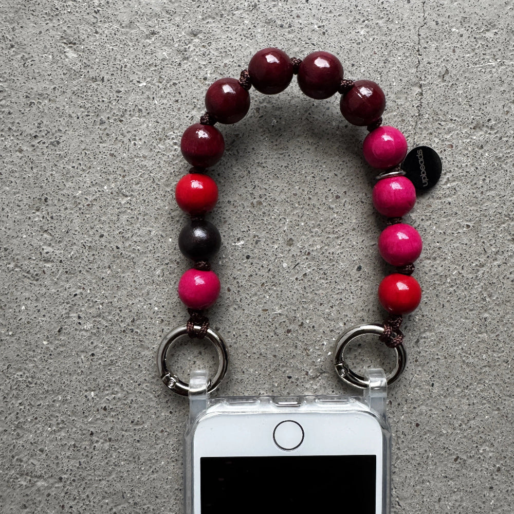 A beaded upbeads phone strap with dark red, bright pink, red and dark brown  wooden signature beads, featuring two silver rings at the bottom and shown holding a white smartphone on a gray concrete surface.