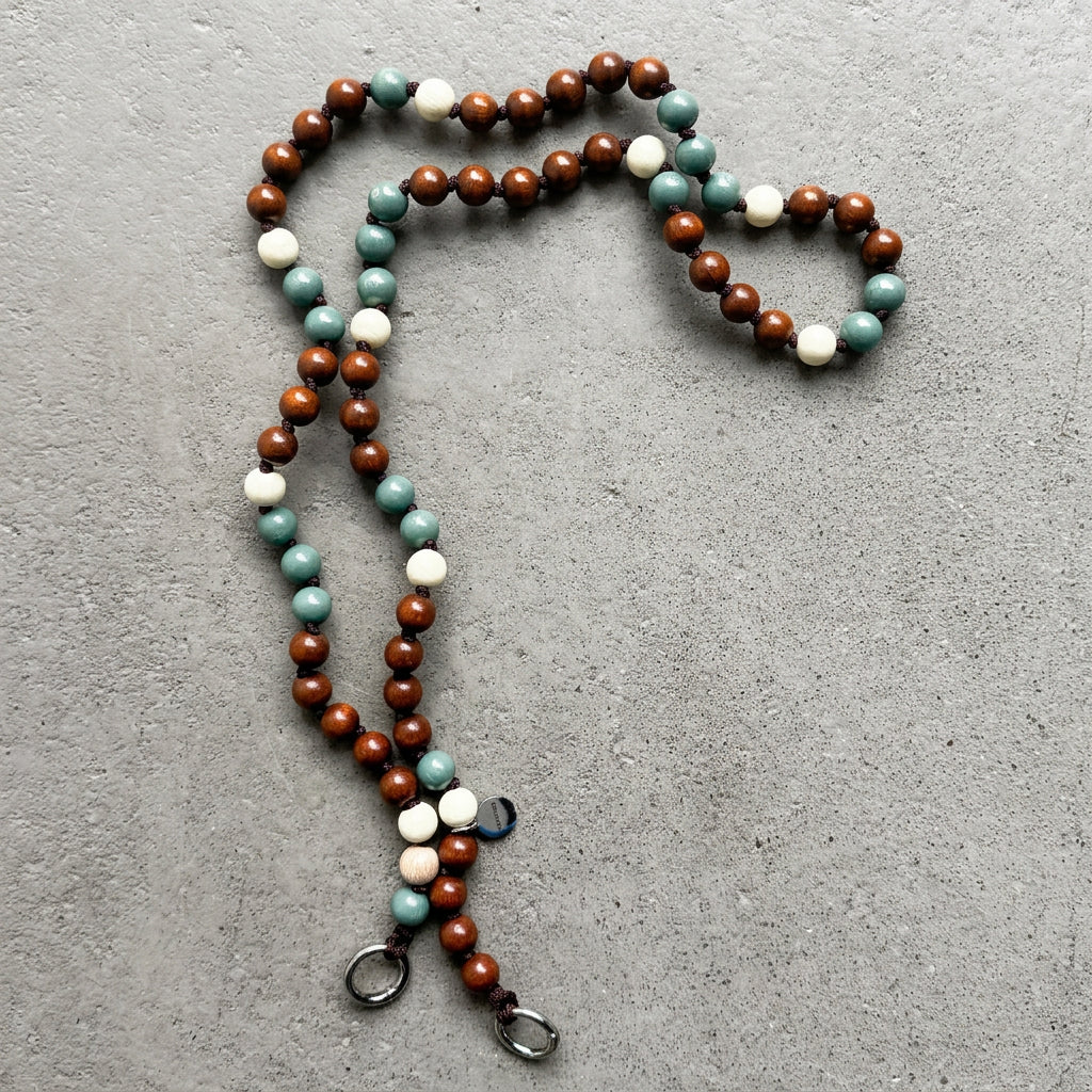 A beaded crossbody cellphone chain with alternating mocca brown, sage green, and creme white round FSC wooden beads, featuring two silver metal carabiner ends, laid out on a textured gray surface.