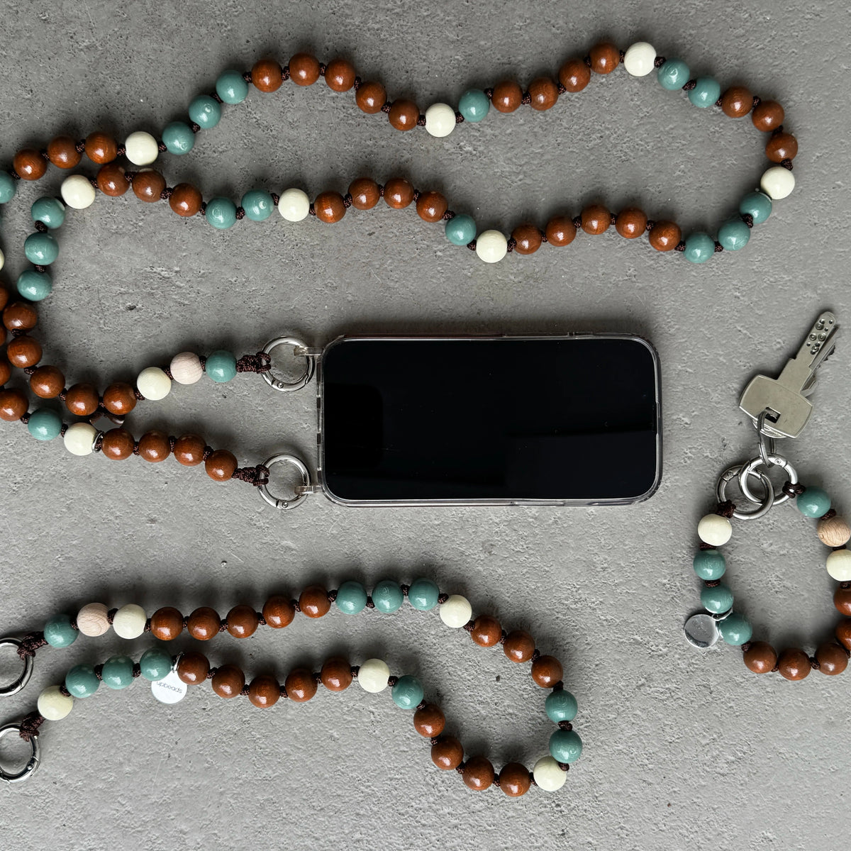 upbeads as phone strap keychain set in 3 different sizes with brown, sage, and cream FSC wooden beads, shown laid out on a gray textured surface alongside a smartphone, keys.