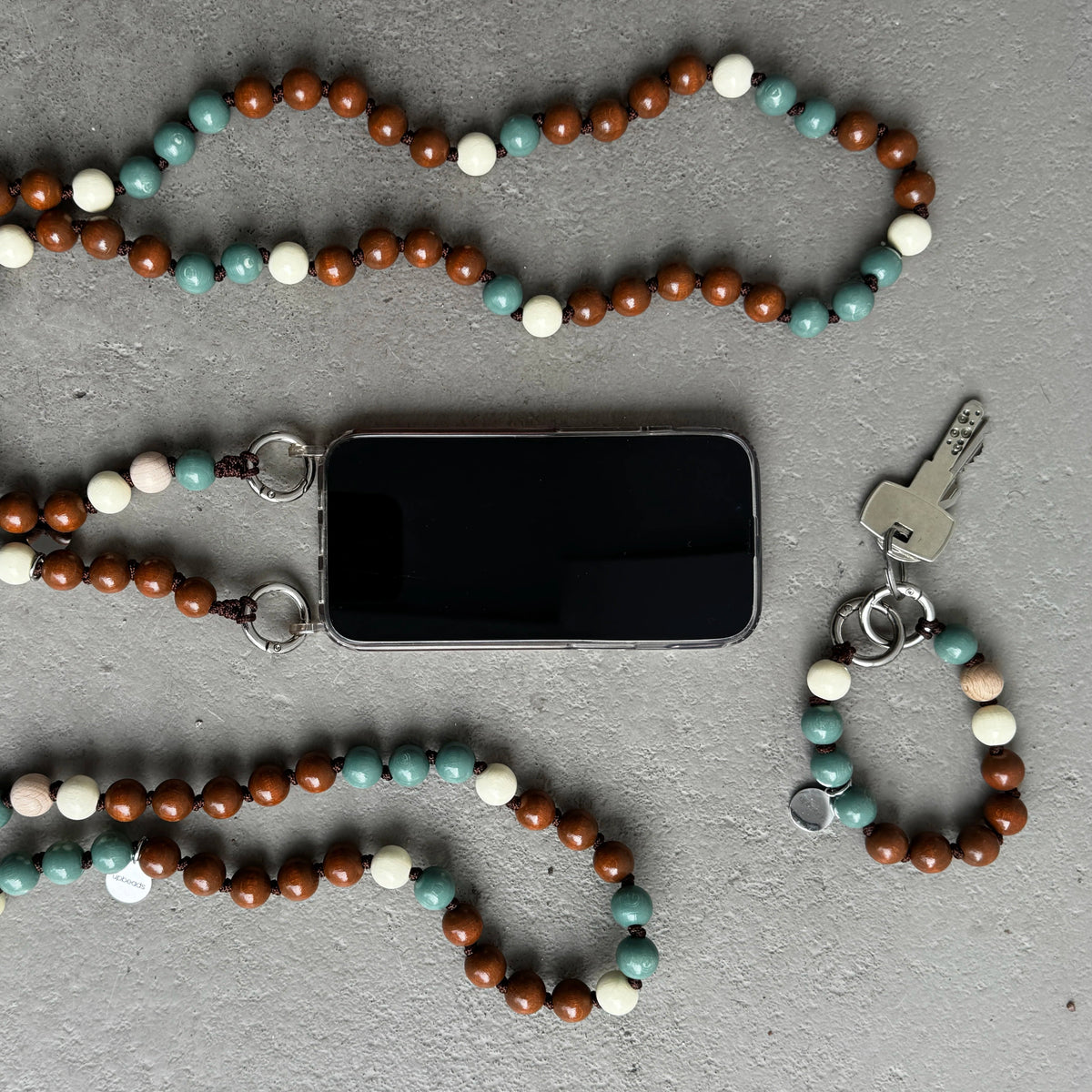 A beaded phone strap keychain set in 3 different sizes with brown, sage, and cream wooden beads, shown laid out on a gray textured surface alongside a smartphone, keys, and metal key rings.