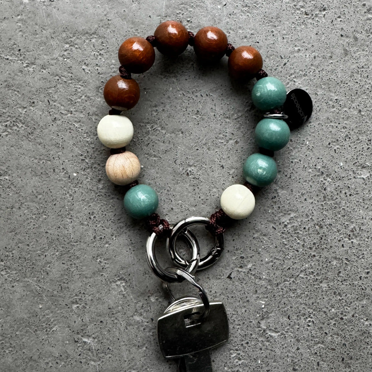 An upbeads keychain bracelet with brown, sage, and creme white wooden beads, attached to silver key rings holding a metal key, displayed on a textured gray concrete surface.