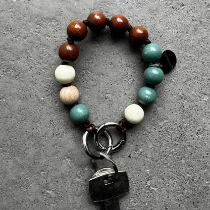 An upbeads keychain bracelet with brown, sage, and creme white wooden beads, attached to silver key rings holding a metal key, displayed on a textured gray concrete surface.