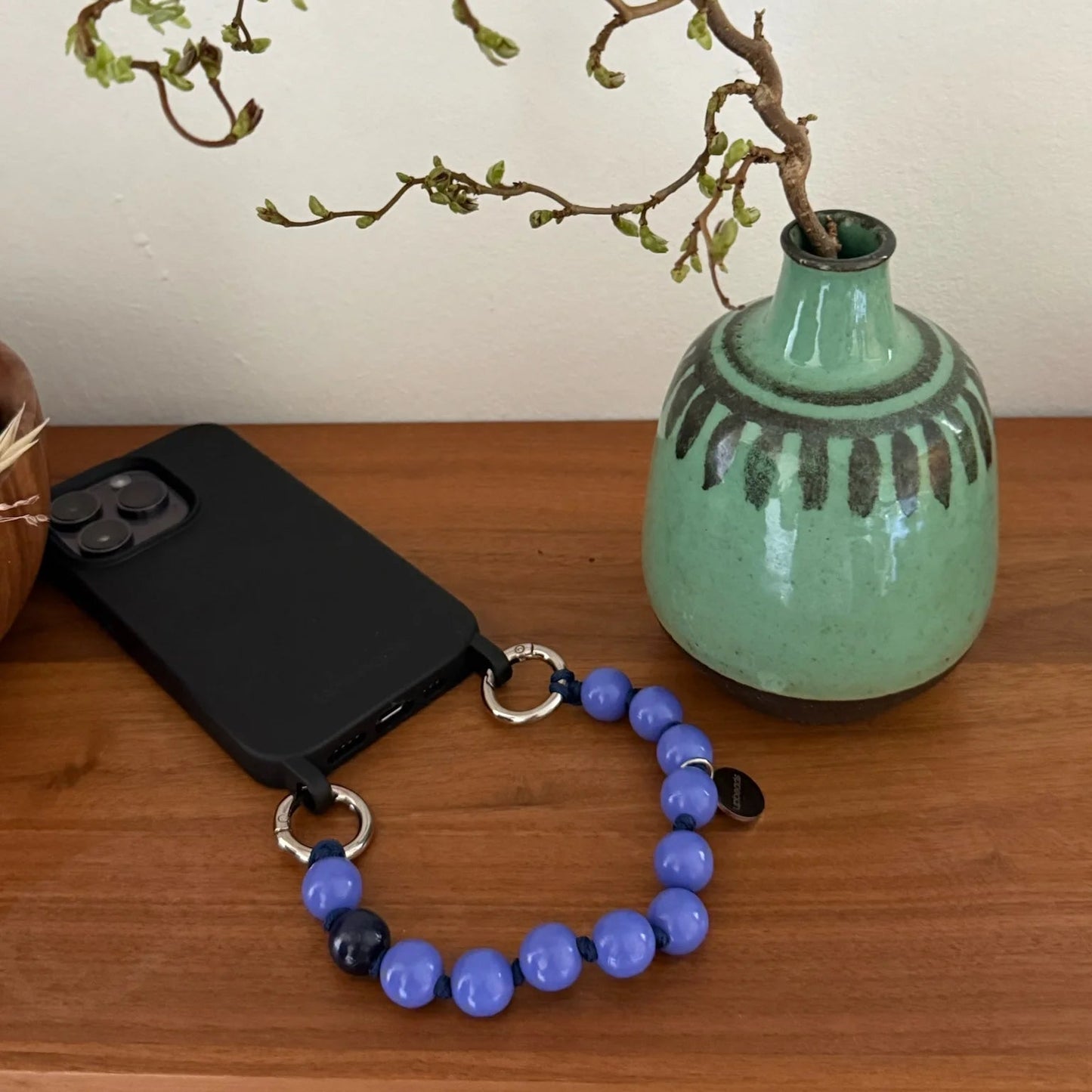 Green vase with branches, black phone case, and purple blue wooden beaded upbeads bracelet as a phone chain on a wooden surface.